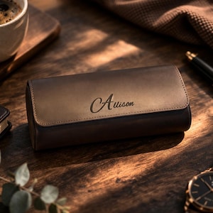 May include: A brown leather watch case with the name "Alison" engraved on the top. The case is rectangular and has a flap closure. A wristwatch with a brown leather strap and a black face is visible. A cup of coffee and a pen are also in the image.