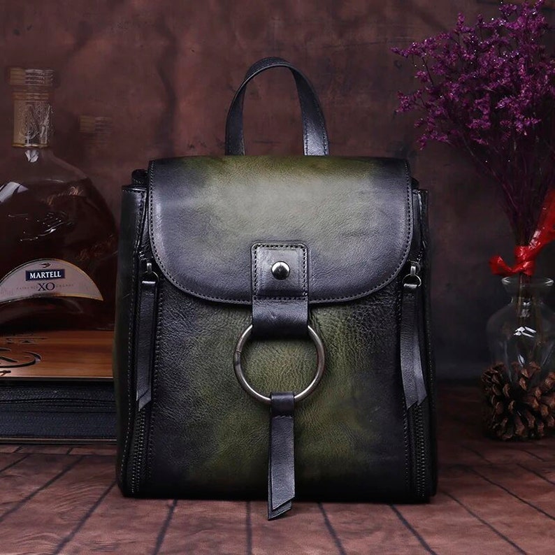 Handcrafted Green leather backpack for women, featuring a classic design with a large metal ring, front strap closure, and side zipper details, showcased in an artistic setting with purple accents