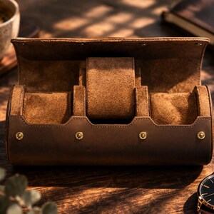 May include: A brown leather watch case with a suede interior, open to display three watch compartments. The case has gold-colored rivets and a matching brown leather strap watch is visible. The case is on a wooden surface.