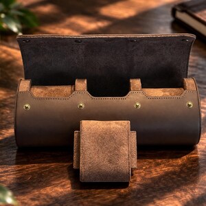 May include: A brown leather watch roll with a suede interior and a matching watch holder. The roll is cylindrical with a flap closure secured by three snaps. Gold-colored rivets accent the design. A notebook is visible in the background.