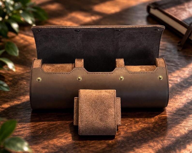 May include: A brown leather watch roll with a suede interior and a matching watch holder. The roll is cylindrical with a flap closure secured by three snaps. Gold-colored rivets accent the design. A notebook is visible in the background.