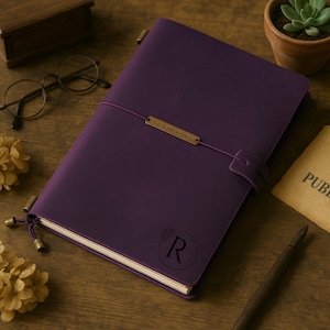 May include: A deep purple leather journal with a strap closure and a decorative metal accent. The journal features an embossed floral design with the letter "R" and a small gold-colored metal plate with text. The journal is on a wooden surface.