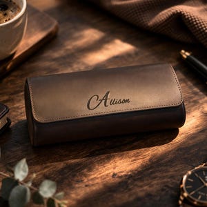 May include: A brown leather watch case with the name "Alison" engraved on the top. The case is rectangular and has a flap closure. A wristwatch with a brown leather strap and a black face is visible. A cup of coffee and a pen are also in the image.