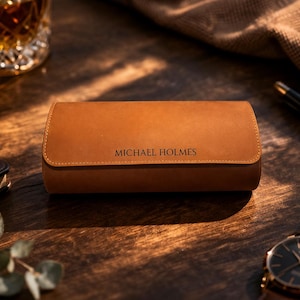 May include: A brown leather watch case with the name "MICHAEL HOLMES" embossed on the front. The case is rectangular with rounded edges and a stitched border. A wristwatch with a black face and gold accents is visible.