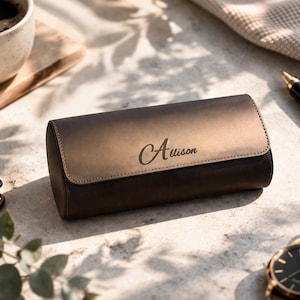 May include: A brown leather watch case with the name "Auison" engraved on the front. The case is cylindrical with a flap closure. A wristwatch with a brown leather band and a black face is visible.