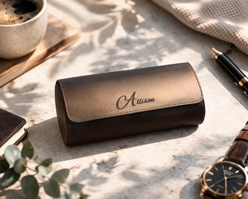 May include: A brown leather watch case with the name "Auison" engraved on the front. The case is cylindrical with a flap closure. A wristwatch with a brown leather band and a black face is visible.