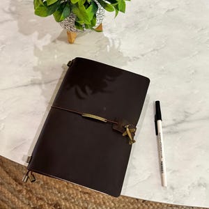 Op de afbeelding: Een donkerbruin leren notitieboek met een decoratieve vliegtuigbedel en een bijpassende pen. Het notitieboek is gesloten met een elastiek en een metalen sluiting. De pen is wit met zwarte inkt.