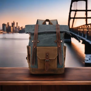 May include: A grey-green canvas backpack with brown leather straps and a front pocket. The bag has a roll-top closure and a brown base. The background features a blurred cityscape and a bridge, creating a travel theme.