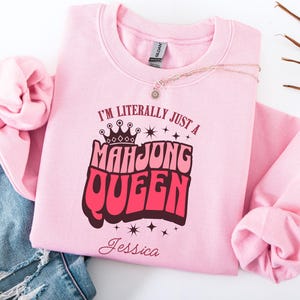May include: A light pink sweatshirt with the text "I'M LITERALLY JUST A MAH JONG QUEEN" in a retro font, with a crown graphic. The name "Jessica" is below the text. A gold necklace and blue jeans are also in the image.