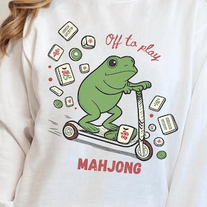 May include: White sweatshirt with a green frog riding a scooter, surrounded by Mahjong tiles. The text "Off to play" is above the frog, and "MAHJONG" is below. The sweatshirt is labeled "Ash" in a blue banner.