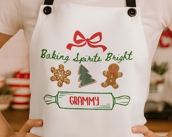 Puede incluir: Delantal blanco con correas negras. Presenta el texto "Baking Spirits Bright" y un lazo rojo. El delantal tiene ilustraciones verdes y rojas de un copo de nieve, un árbol de Navidad y un hombre de jengibre. La palabra "Grammy" está impresa debajo.