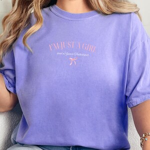 Pu&ograve; includere: Una maglietta color lavanda con il testo "I'm just a girl (and a Speech Pathologist)" in lettere rosa con un fiocco rosa.