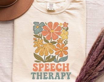 May include: Cream-colored t-shirt with a retro floral design in orange, yellow, blue, and peach. The words "SPEECH THERAPY" are printed in a matching retro font. The shirt is a Comfort Colors XL-TG-EG.