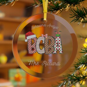 May include: A clear, round Christmas ornament with a gold ribbon. The ornament features the text "Behavioral Analyst" and "BCBA" in festive patterns, including a Santa hat and elf legs. The name "Mrs. Ricciardi" is also displayed.