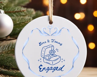 May include: White ceramic engagement ornament with blue text and design. The ornament features the names "Gina & Tommy", a ring in a box, the word "Engaged", and the year "2025". A twine loop is at the top for hanging.