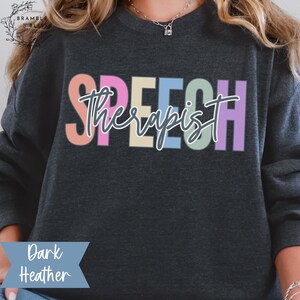 May include: Dark heather gray sweatshirt with the word "SPEECH" in large, colorful letters across the chest. The word "Therapist" is written in a cursive font over the word "SPEECH". The words "Dark Heather" are printed in a blue banner at the bottom.