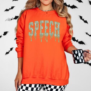 May include: An orange sweatshirt with the word "SPEECH" in a dripping, textured font. The sweatshirt is paired with black and white checkered pants. A black mug with white ghost designs is held in the hand.