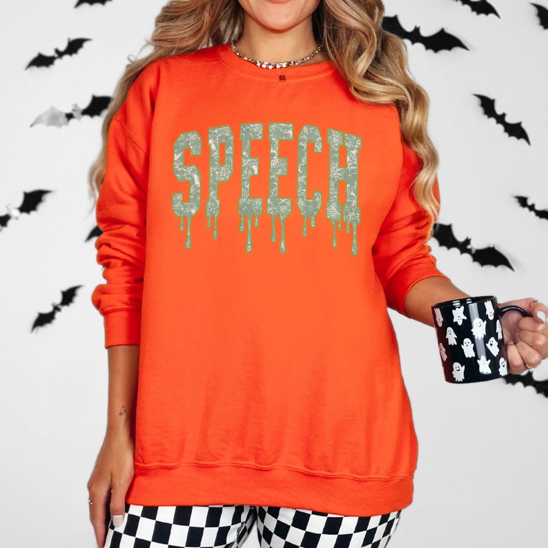 May include: An orange sweatshirt with the word "SPEECH" in a dripping, textured font. The sweatshirt is paired with black and white checkered pants. A black mug with white ghost designs is held in the hand.
