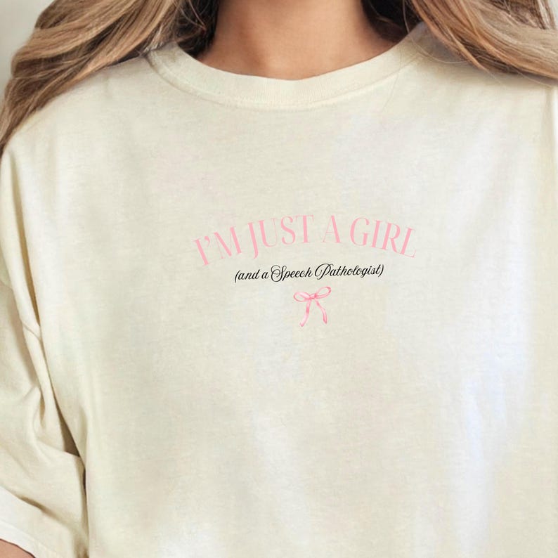 Pu&ograve; includere: Una maglietta bianca con il testo "I'm just a girl (and a Speech Pathologist)" in nero con un fiocco rosa sotto.