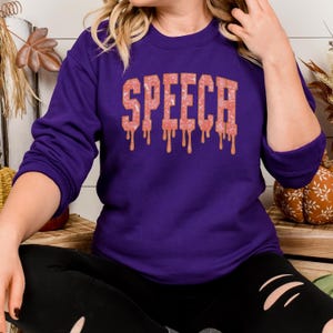 May include: A purple sweatshirt with the word "SPEECH" in a pink, textured font with dripping accents. The sweatshirt is a solid color, and the text is the focal point. The image is of a person wearing the sweatshirt.