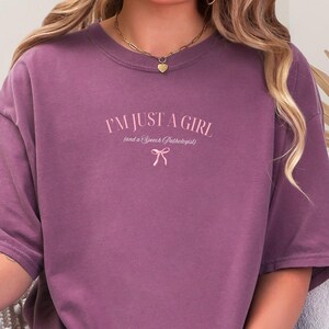 Pu&ograve; includere: Una maglietta viola con il testo "I'm just a girl (and a speech pathologist)" in lettere rosa con un fiocco rosa sotto.