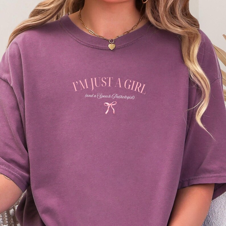Pu&ograve; includere: Una maglietta viola con il testo "I'm just a girl (and a speech pathologist)" in lettere rosa con un fiocco rosa sotto.