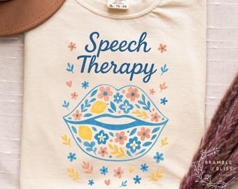 May include: Cream-colored t-shirt with the words "Speech Therapy" in blue script above a blue outline of lips filled with flowers and lemons. The shirt has a Comfort Colors tag and is decorated with small flowers and leaves.