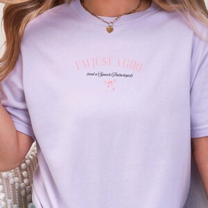 Pu&ograve; includere: Una maglietta color lavanda con il testo "I'm just a girl (and a Speech Pathologist)" in nero con un fiocco rosa sotto.