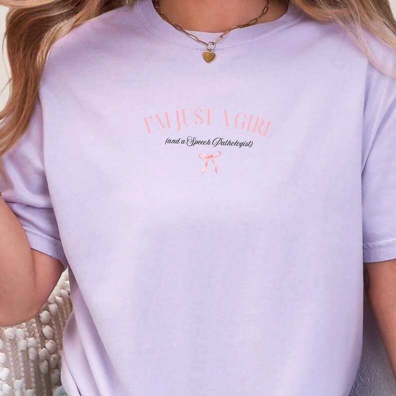 Pu&ograve; includere: Una maglietta color lavanda con il testo "I'm just a girl (and a Speech Pathologist)" in nero con un fiocco rosa sotto.