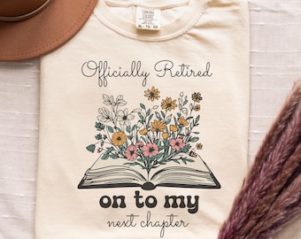 May include: A cream-coloured t-shirt with the text "Officially Retired on to my next chapter" and a floral illustration of a book with flowers on top.