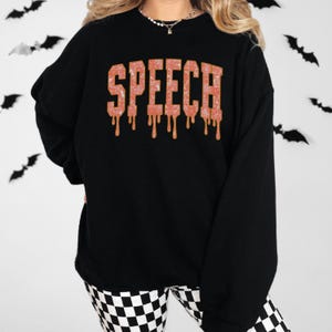 May include: Black sweatshirt with the word "SPEECH" in orange, dripping letters. The sweatshirt is paired with black and white checkered pants. Bats are in the background.