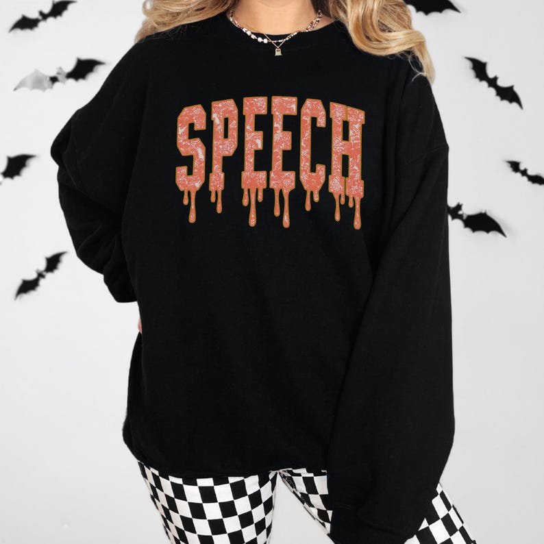 May include: Black sweatshirt with the word "SPEECH" in orange, dripping letters. The sweatshirt is paired with black and white checkered pants. Bats are in the background.