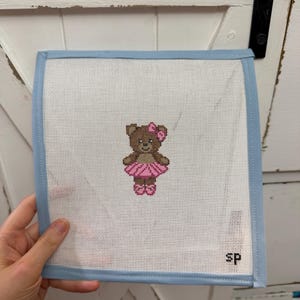 May include: Square needlepoint canvas featuring a brown teddy bear wearing a pink tutu and bow. The canvas has a white background with a light blue border. The initials "sp" are in the bottom right corner.