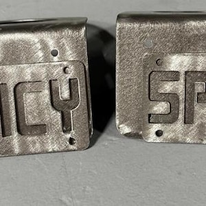 May include: Two rectangular metal brackets with a brushed silver finish, each featuring a flat front plate with the word SPICY cut out in bold, block capital letters. Each plate includes four small circular mounting holes at the corners.