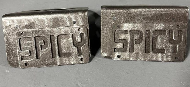 May include: Two rectangular metal brackets with a brushed silver finish, each featuring a flat front plate with the word SPICY cut out in bold, block capital letters. Each plate includes four small circular mounting holes at the corners.