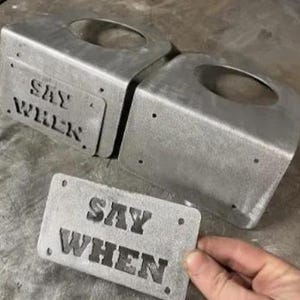 May include: Two silver-colored metal brackets with circular cutouts and a rectangular metal plate, all featuring the embossed text SAY WHEN. The components have a raw, industrial finish with visible screw holes on the plate and bracket faces.