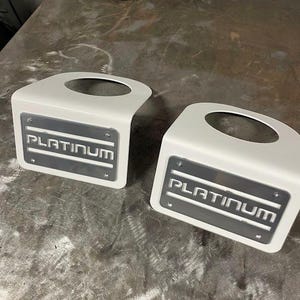 May include: Two white metal automotive parts with a curved design and a circular opening. Each part has a rectangular plate with the word "PLATINUM" in a dark gray, industrial font. The parts are on a metallic surface.