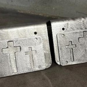 May include: Two rectangular silver-colored metal plates with a bent top edge. Each plate features a cutout design of three crosses arranged horizontally in the center. There are small circular holes punched near the corners of each metal piece.