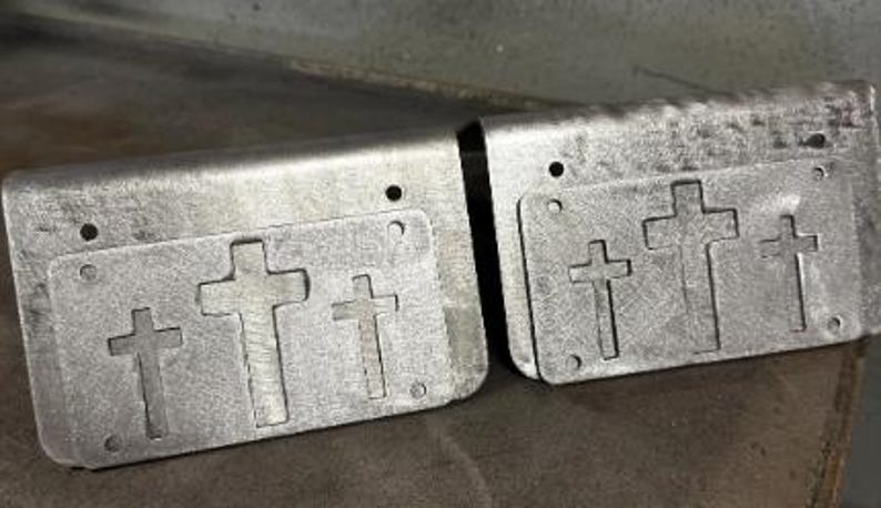 May include: Two rectangular silver-colored metal plates with a bent top edge. Each plate features a cutout design of three crosses arranged horizontally in the center. There are small circular holes punched near the corners of each metal piece.