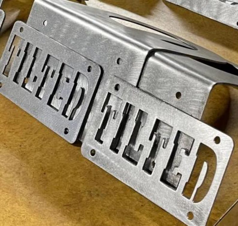 May include: A silver-colored metal license plate frame featuring the word TILTED cut out in a bold, block font. The frame has a brushed finish and includes four circular mounting holes, two at the top and two at the bottom, for secure attachment.