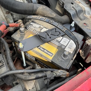 May include: A black metal car battery tie-down bracket featuring the cutout text MK3. The bracket is installed over a black and yellow automotive battery with a 6-year warranty label, secured by a silver metal bolt and nut within an engine bay.