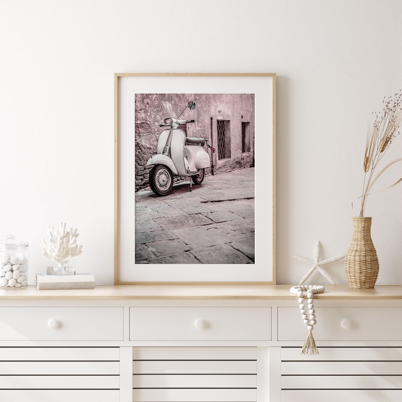 May include: A framed photograph of a white vintage scooter parked in a narrow, cobblestone alleyway. The scooter is in focus, while the background is blurred and faded in a soft pink hue.