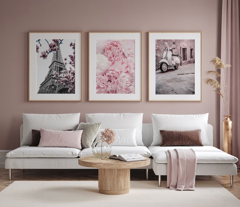 May include: Three framed prints on a pink wall above a white couch. The prints feature a black and white photo of the Eiffel Tower, a close-up of pink peonies, and a vintage scooter parked in a cobblestone street.