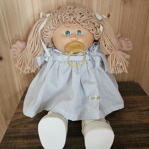 Op de afbeelding: Een vintage pop met blond garen haar in staartjes, gekleed in een blauw-wit geruite jurk en witte schoenen. De pop heeft een fopspeen en groene ogen. De pop zit op een houten oppervlak.