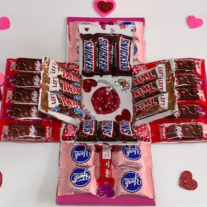 Może przedstawiać: Różowe i białe pudełko eksplodujące wypełnione czekoladowymi batonikami. Pudełko jest ozdobione sercami i napisami "LEFT" i "Twix". Batoniki czekoladowe to Snickers, Twix i York Peppermint Patties.