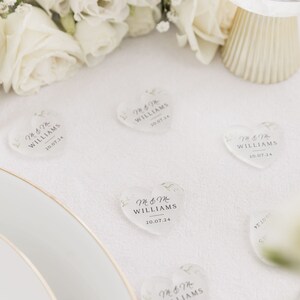 May include: Clear heart-shaped acrylic wedding favors with the text "Mr & Mrs Williams 20.07.24" printed on them. The favors are scattered on a white tablecloth.