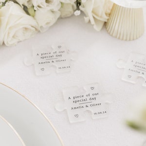 May include: Three clear acrylic puzzle pieces with the text "A piece of our special day Amelia & Oliver" and the date "16.08.24" printed on them. The puzzle pieces are on a white tablecloth.