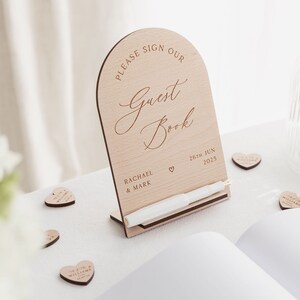 May include: A wooden arch-shaped sign with the text "Please sign our Guest Book" in cursive script. The sign also includes the names "Rachael & Mark" and the date "26th Jun 2025".