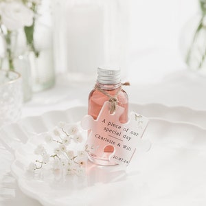 May include: A small clear bottle with a pink liquid and a white puzzle piece tag that says "A piece of our special day Charlotte & Will 20.07.24". The bottle is sitting on a white plate with white flowers.