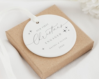 Może przedstawiać: Okrągła biała ceramiczna ozdoba z białą wstążką, zawierająca czarny napis OUR FIRST Christmas ENGAGED, RACHEL & OLIVER oraz 2026, ozdobiona małymi symbolami gwiazdek, spoczywająca na kwadratowym brązowym kartonowym pudełku prezentowym.